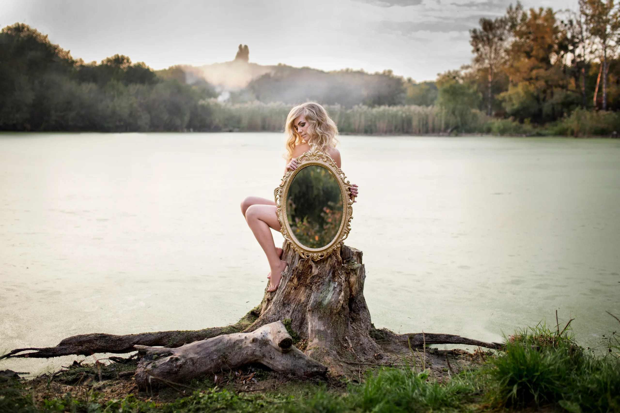 Blonde naked woman holds a mirror sitting before a lake covered. Glorious Mystery Meditation