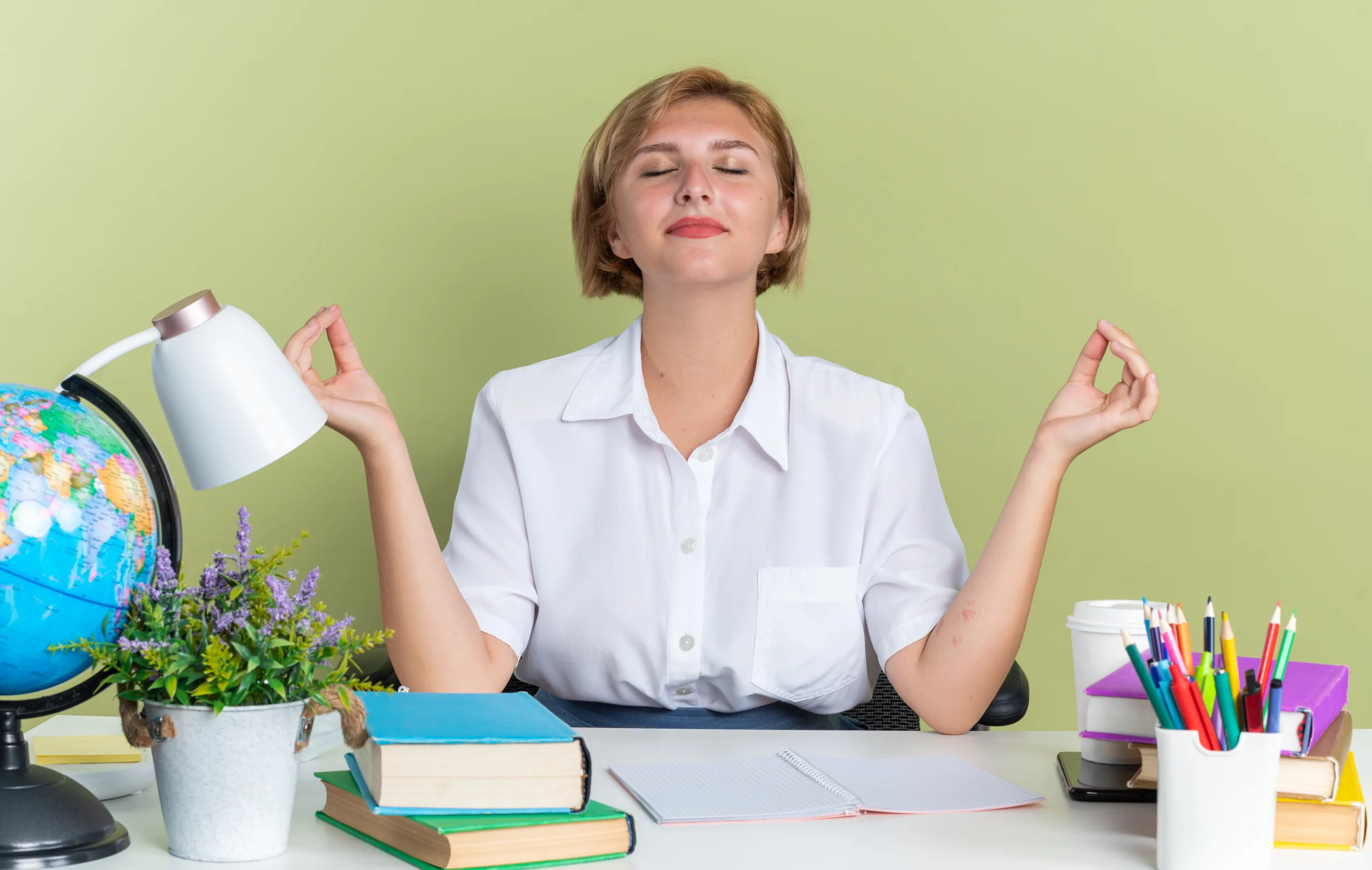 peaceful young blonde teacher sitting at desk with school tools meditating with closed eyes isolated on olive green background